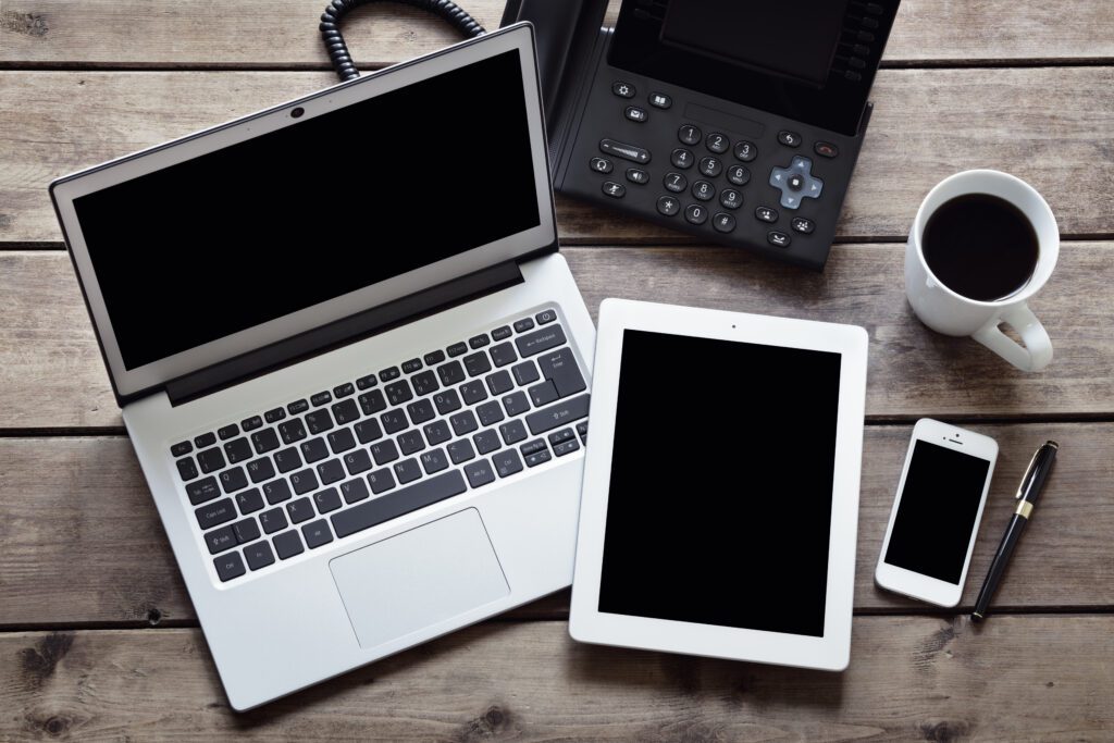 Open laptop with Ip desk phone, white tablet and smartphone on desk from above. Comparison of virtual and business phone options.